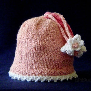 Puede incluir: Un gorro de punto rosa con una flor blanca de ganchillo y un borde blanco de ganchillo alrededor del borde.