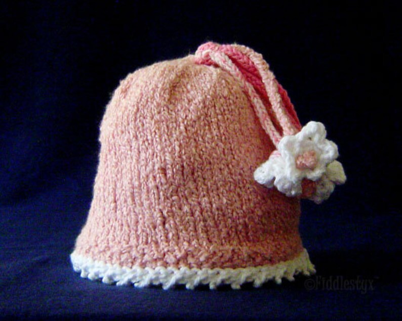 Puede incluir: Un gorro de punto rosa con una flor blanca de ganchillo y un borde blanco de ganchillo alrededor del borde.