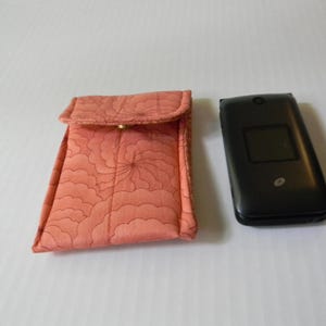 Peut inclure: Un étui en tissu corail avec un motif floral et un téléphone à clapet noir. L'étui a une fermeture à rabat avec un bouton doré. Le téléphone est fermé et possède un petit écran.