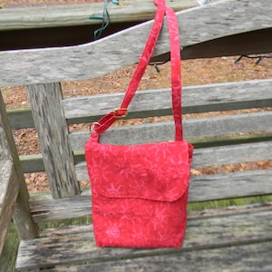 Può includere: Una borsa a tracolla in tessuto rosso con chiusura a lembo e una lunga tracolla regolabile. La borsa ha un design a sole vorticoso.