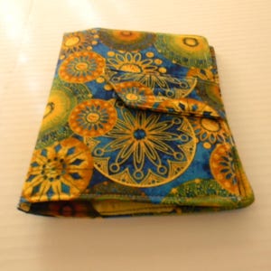 Op de afbeelding: Een kleurrijke portemonnee met een bloemenpatroon in blauw, geel en groen. De portemonnee heeft een klepsluiting en is gemaakt van stof. Het ontwerp heeft cirkelvormige en stervormige motieven.