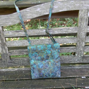 May include: A rectangular crossbody bag featuring a vibrant batik-style fabric pattern in shades of teal, blue, and purple. The bag has a fold-over flap closure and a matching adjustable fabric shoulder strap, showcasing a repeating circular motif.