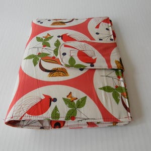 May include: A folded fabric case with a flap closure. The case features a repeating pattern of red cardinals, green leaves, and orange butterflies on a white background with red circular accents. The case is likely for storing items.
