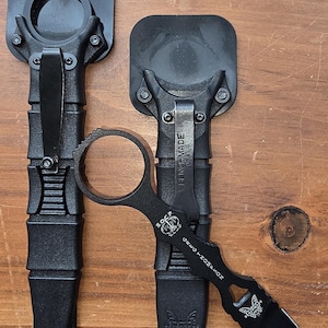 Peut inclure: Deux couteaux tactiques noirs avec un logo papillon et le texte "Pinchmade USA" sur les étuis. Les couteaux ont une poignée en anneau et une lame noire.