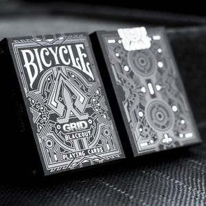 May include: Two decks of Bicycle playing cards with a black and silver design. The front of the deck features the text "Bicycle" and "Grid Blackout Playing Cards". The back of the deck features a geometric pattern.