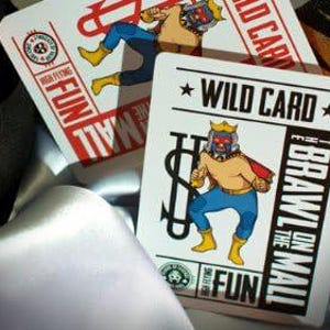 Op de afbeelding: Een witte speelkaart met een rode en zwarte illustratie van een worstelaar met een dollarteken en de tekst "WILD CARD" en "I BRAWL ON THE MALL".