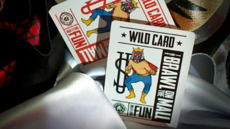 Op de afbeelding: Een witte speelkaart met een rode en zwarte illustratie van een worstelaar met een dollarteken en de tekst "WILD CARD" en "I BRAWL ON THE MALL".