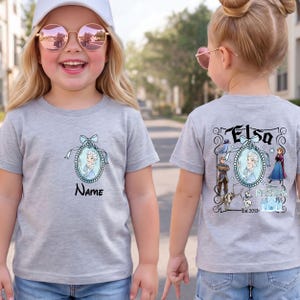 May include: A heather grey t-shirt featuring a front graphic of Elsa in a blue oval frame with a bow and the word Name. The back graphic displays Elsa, Anna, Kristoff, Olaf, Sven, and the Frozen logo with the text Elsa and Est. 2013.