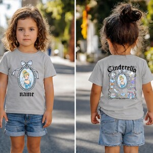 Puede incluir: Camiseta gris claro con un dise&ntilde;o de Cenicienta en la parte delantera y trasera. El dise&ntilde;o frontal incluye un retrato de Cenicienta y la palabra "name". El dise&ntilde;o trasero incluye la palabra "Cenicienta" e ilustraciones relacionadas.