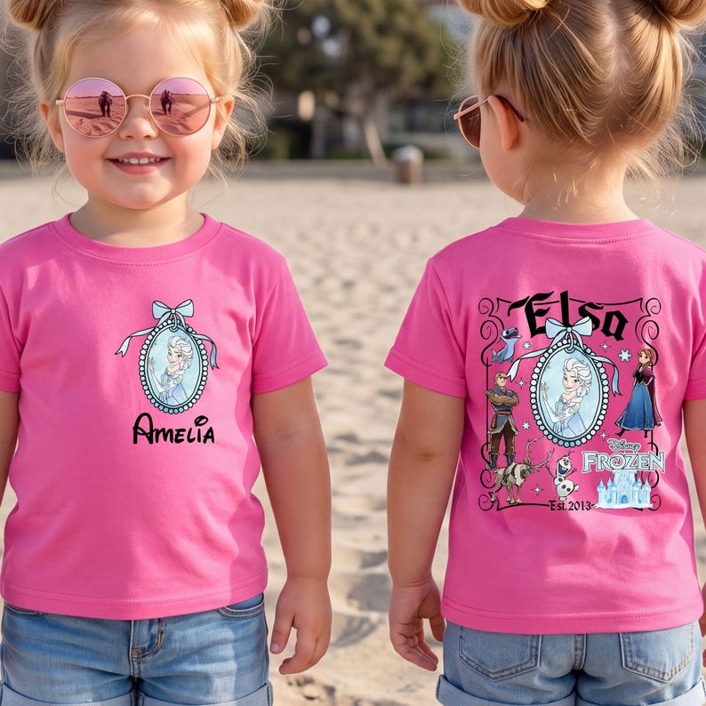 May include: A bright pink short-sleeved t-shirt featuring a front graphic of Elsa in an oval frame with the name Amelia below. The back displays a larger Frozen-themed graphic with Elsa, Anna, Kristoff, Sven, Olaf, and the Disney Frozen logo.