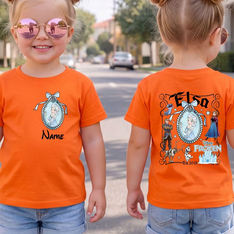 May include: An orange t-shirt featuring a front graphic of Elsa in a blue oval frame with a bow and the word Name. The back graphic displays Elsa, Anna, Kristoff, Olaf, Sven, and a castle with the text Elsa, Disney Frozen, and Est. 2013.