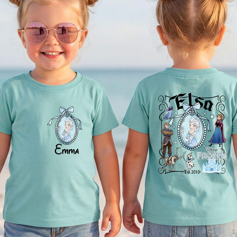 May include: A light teal t-shirt featuring a small oval frame graphic with Elsa on the front and the name Emma. The back displays a large Frozen-themed graphic with Elsa, Anna, Kristoff, Sven, Olaf, and the text Elsa, Disney Frozen, and Est. 2013.