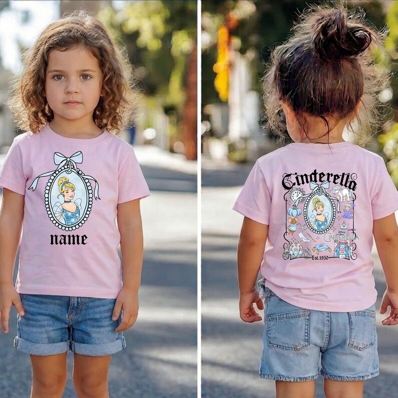 Puede incluir: Una camiseta rosa con un dise&ntilde;o de Cenicienta en la parte delantera y trasera. El frente muestra un retrato de Cenicienta y la palabra "name". La parte trasera presenta la palabra "Cenicienta" e ilustraciones relacionadas.
