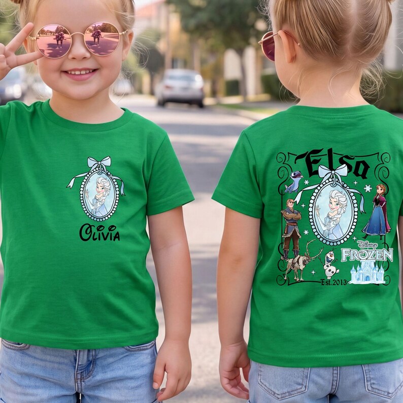 May include: A bright green t-shirt featuring a small oval cameo of Elsa on the front chest with the name Olivia below. The back displays a large graphic with Elsa, Anna, Kristoff, Sven, Olaf, and the Frozen logo with the text Est. 2013.