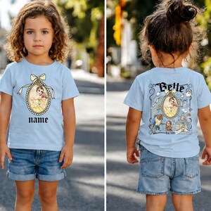 Puede incluir: Camiseta azul claro con un retrato de princesa en vestido amarillo y un espacio para un nombre. La parte trasera muestra el nombre "Belle" sobre un retrato, rodeado de ilustraciones de personajes y un castillo. El texto "Est. 1991" est&aacute; impreso debajo.