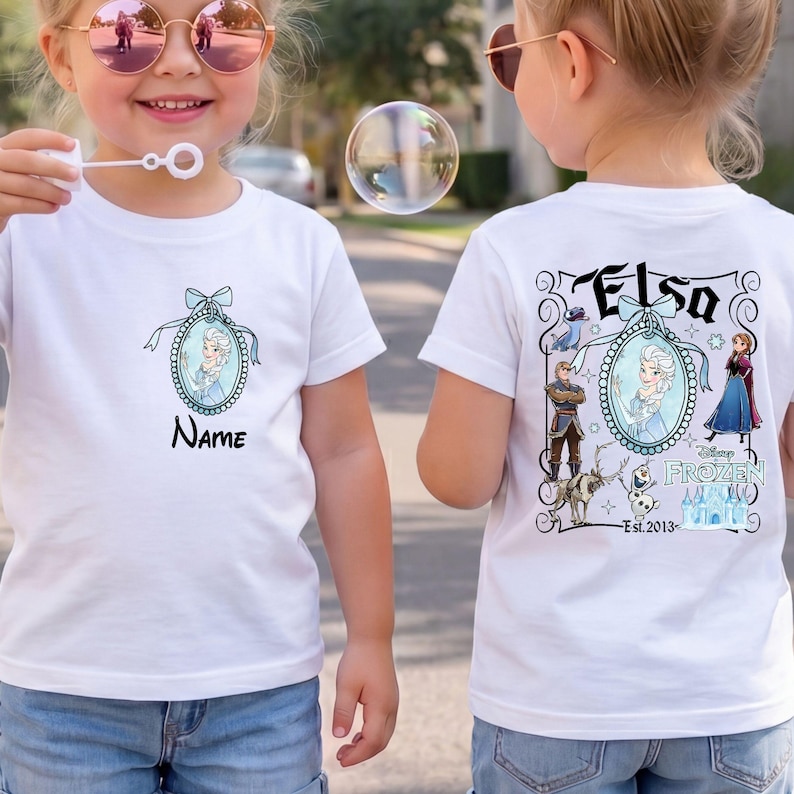 May include: A white short-sleeved t-shirt featuring a small oval graphic of Elsa on the front chest with the text Name below it. The back of the shirt displays a larger Frozen-themed graphic with Elsa, Anna, Kristoff, Sven, Olaf, and a castle.