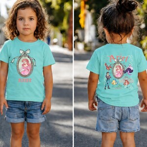 Puede incluir: Una camiseta turquesa con una imagen de dibujos animados de una princesa con un vestido rosa dentro de un marco ovalado con un lazo. El nombre "Name" est&aacute; impreso debajo. La parte posterior de la camiseta tiene la imagen de la princesa y otros personajes.