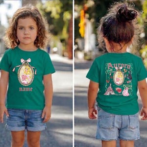 Puede incluir: Una camiseta verde azulado con un dise&ntilde;o de princesa y un campo de nombre. El dise&ntilde;o frontal incluye una princesa con un vestido rosa dentro de un marco ovalado con un lazo. La parte posterior de la camiseta tiene el texto "Aurora Sleeping Beauty" con gr&aacute;ficos adicionales.