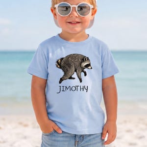 May include: A light blue short-sleeved t-shirt featuring a detailed illustration of a raccoon in shades of gray and brown. Below the raccoon, the name JIMOTHY is printed in a simple, dark, hand-drawn style font across the center of the chest.
