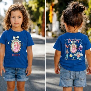 Puede incluir: Una camiseta azul con una imagen de dibujos animados de una princesa con un vestido rosa dentro de un marco ovalado con un lazo. La palabra "name" est&aacute; debajo. La parte posterior de la camiseta tiene el nombre "Ariel" y otros personajes de la pel&iacute;cula.