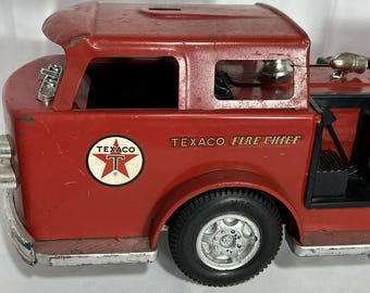 Op de afbeelding: Rode speelgoedbrandweerwagen met zwarte wielen en een Texaco Fire Chief logo aan de zijkant. De wagen heeft een ladder aan de achterkant en een bel bovenop.