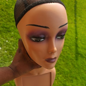 May include: A tan mannequin head with painted purple eyeshadow, dark eyebrows, and dark lipstick. A black mesh wig cap is on the head. The mannequin is held by a dark-skinned hand.