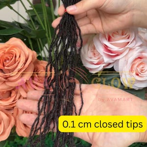 Könnte beinhalten: Ein Bündel dunkelbrauner Dreadlocks mit 0,1 cm geschlossenen Spitzen wird vor einem Hintergrund aus pfirsichfarbenen und rosa Rosen gehalten. Die Dreadlocks sind dünn und strukturiert, mit einem natürlichen Aussehen.