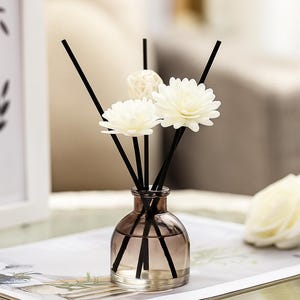 Puede incluir: Un difusor de caña decorativo con una botella de vidrio transparente y cañas negras. La botella contiene un líquido marrón claro y está adornada con decoraciones blancas en forma de flor. El difusor está sobre una revista.