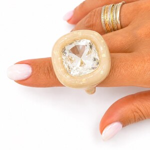 May include: A chunky ring featuring a large, clear, cushion-cut faceted stone set within a smooth, beige, resin-like frame. The band is thick and rounded, complementing the prominent central gemstone. Additional stacked metallic rings are visible.