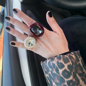 May include: Two chunky rings featuring large, faceted, rectangular-cut stones. One ring has a deep burgundy setting with a dark gray stone, while the other has a cream-colored setting with a clear, sparkling stone, both worn on the fingers.