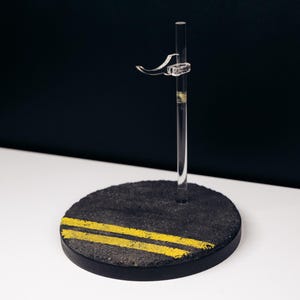 May include: A clear acrylic display stand with a black, circular base featuring two yellow stripes, resembling a road. The stand has a clear vertical support and a curved hook for holding an item. The background is black and white.