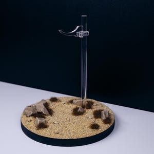 May include: A clear acrylic display stand with a circular base featuring sand, rocks, and tufts of grass. The stand has a vertical support and a curved arm for holding an item. The base is black-edged.