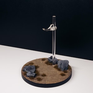 May include: A clear acrylic display stand with a circular base featuring a textured brown surface, small rocks, and a vertical support with a curved arm. The stand is designed to hold a small object.