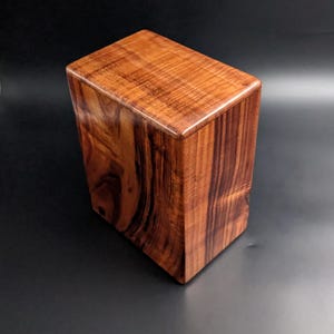 May include: A rectangular wooden box with a polished finish, showcasing a rich reddish-brown color with prominent dark grain patterns. The box features a smooth, flat top surface and vertical sides, displaying natural wood textures throughout the exterior.