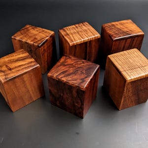 May include: Six wooden cubes with varying grain patterns and rich brown hues are arranged on a dark surface. The cubes have a polished finish, highlighting the natural wood grain. The cubes are a decorative item.
