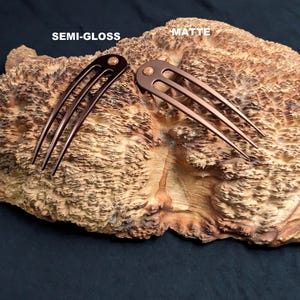 May include: Two bronze hair forks, one semi-gloss and one matte, are displayed on a textured wooden surface. Each fork has three tines and a decorative top. The text "SEMI-GLOSS" and "MATTE" are visible above the forks.