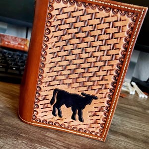 Könnte beinhalten: Ein braunes Lederjournal mit einem Webmuster und einer schwarzen Bullensilhouette. Das Journal hat einen dekorativen Rand und einen genähten Rücken. Der Einband ist heller braun als der Rücken.