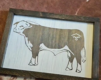 May include: Wooden framed artwork featuring a bull silhouette. The bull is depicted in dark brown against a light background, with a dark brown frame. A small brand is visible on the bull's side. The artwork is displayed on a brown and white cowhide.