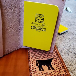 May include: Open leather notebook with a yellow notepad labeled "Rite in the Rain" and "Beef Calving Record Book." The notebook is brown with a decorative border and a black silhouette of a cow.