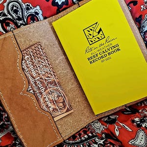 May include: A brown leather notebook cover with a yellow Rite in the Rain Beef Calving Record Book inside. A decorative card with the text "Custom Leatherwork" is in the front pocket. The background is a red paisley pattern.