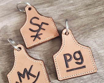 May include: Three leather keychains with metal rings. Each keychain is a light brown color, shaped like a livestock tag, and features a unique black engraved design. The keychains are stitched around the edges.
