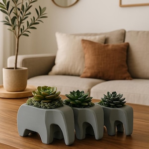 May include: Three gray elephant-shaped planters with succulents. The planters are on a wooden table in front of a beige couch.