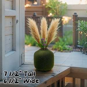 Puede incluir: Un jarrón verde con un diseño en espiral contiene hierba de la pampa seca y pequeñas flores moradas. El jarrón mide aproximadamente 19 cm de alto y 16,5 cm de ancho. El jarrón está sobre una superficie de madera cerca de una puerta blanca.