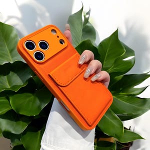 May include: Bright orange phone case with a built-in card holder. The case has a textured surface and a raised section for the camera lenses. The card holder is a flap that secures with a snap. The case is shown against a background of green leaves.