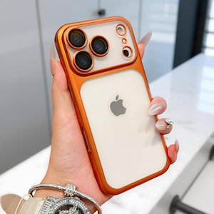 Puede incluir: Una funda de teléfono naranja y transparente con un marco dorado. La funda tiene una parte trasera transparente con el logotipo de Apple y una sección para las lentes de la cámara del teléfono. La funda está sujeta en la mano de una persona.