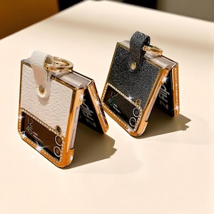 May include: Two open, gold-framed phone cases with a hinged design. One is white with a gold ring, the other is black with a black strap. Both cases have a rhinestone border and the text "Z Flip".