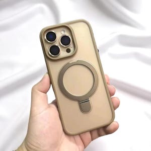 May include: A tan phone case with a circular magnetic ring and a kickstand. The case has a raised edge around the camera lenses. The phone case is held in a hand.
