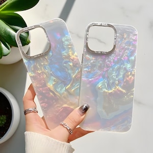 May include: Two iridescent phone cases with a pearlescent finish. The cases have a silver-colored camera frame with the text "OSA BEAN". The cases have a swirling, colorful pattern with hints of pink, blue, and yellow.