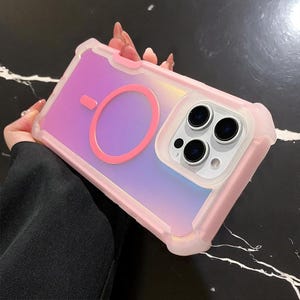 以下が含まれることがあります： ピンクと虹色の携帯電話ケース、マグネットリング付き。ケースは透明な保護バンパーとピンク、紫、青のグラデーションになっています。携帯電話のカメラレンズが見えます。