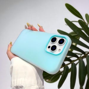 Puede incluir: Una funda de teléfono azul claro con un borde de cámara turquesa. La funda tiene un acabado liso y mate y presenta tres lentes de cámara. La funda del teléfono se sostiene en una mano, con hojas verdes en el fondo.